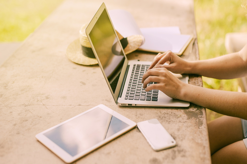 uma mulher com notebook, tablet e celular na mesa, escreve no notebook com folhas ao seu lado, aparentemente estudando online